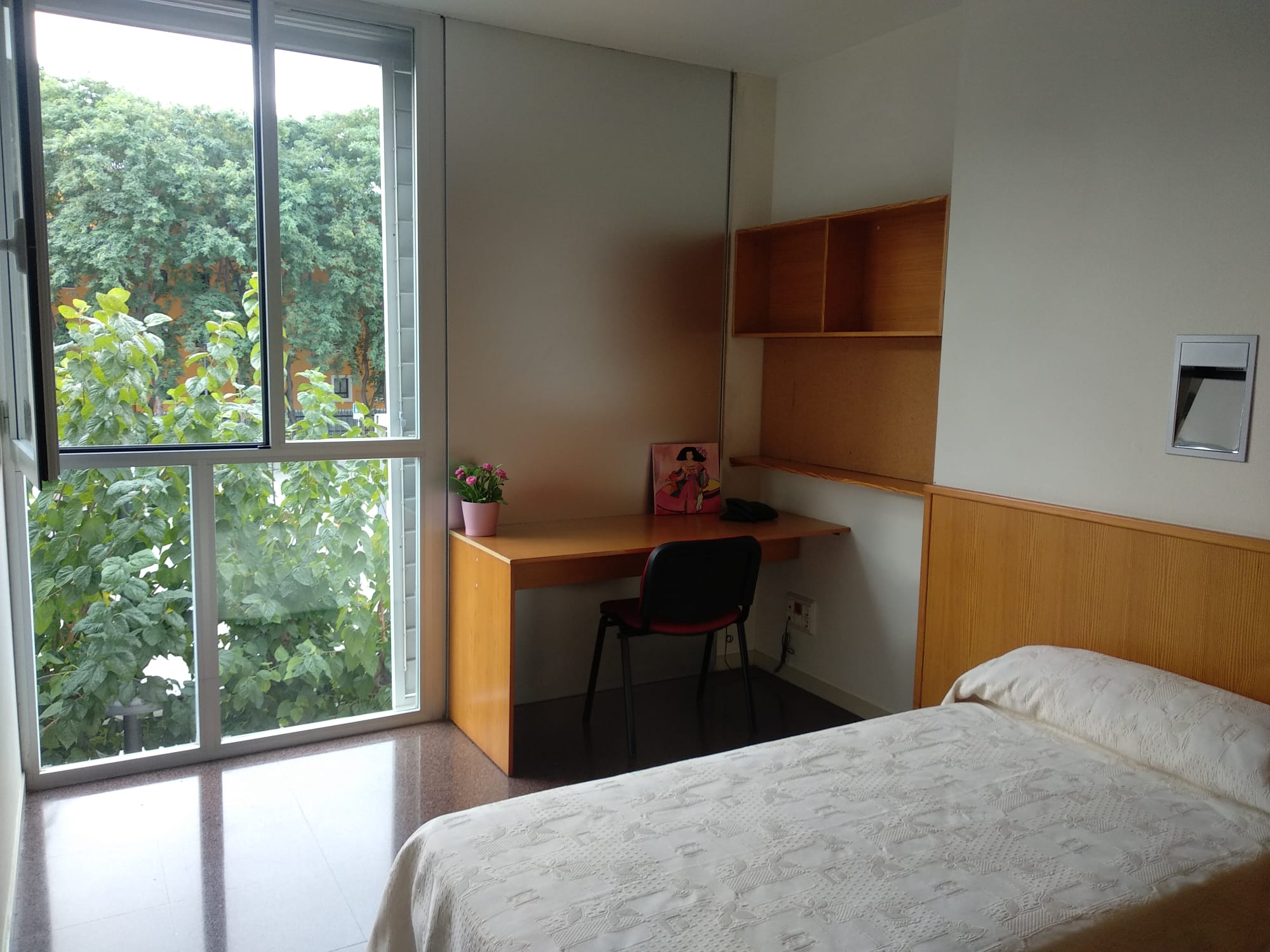 Habitación de residencia de estudiantes con amplio ventanal, cama estantería, mesa y silla